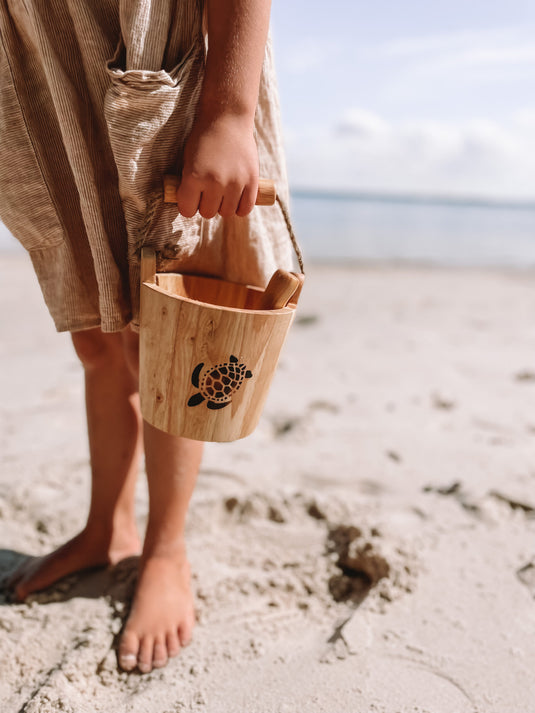 Person holding a wooden bucket with a turtle design on a beach.