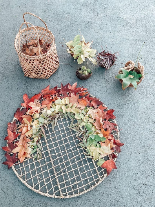 Decorative wreath with autumn leaves on a textured surface with a basket and small plants.