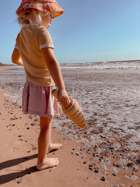 Child on a beach holding a shovel, wearing a hat and pink skirt.
