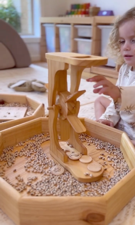 Child playing with a wooden sensory bin filled with small objects in a home setting.