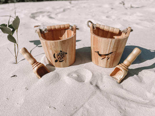 Two wooden buckets and shovels on sandy ground with a plant in the background