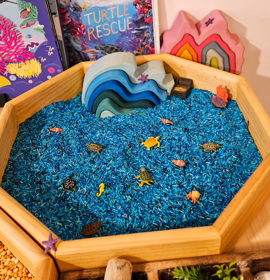 Wooden sensory bin with blue sand, turtle toys, and a book titled 'Turtle Rescue'.
